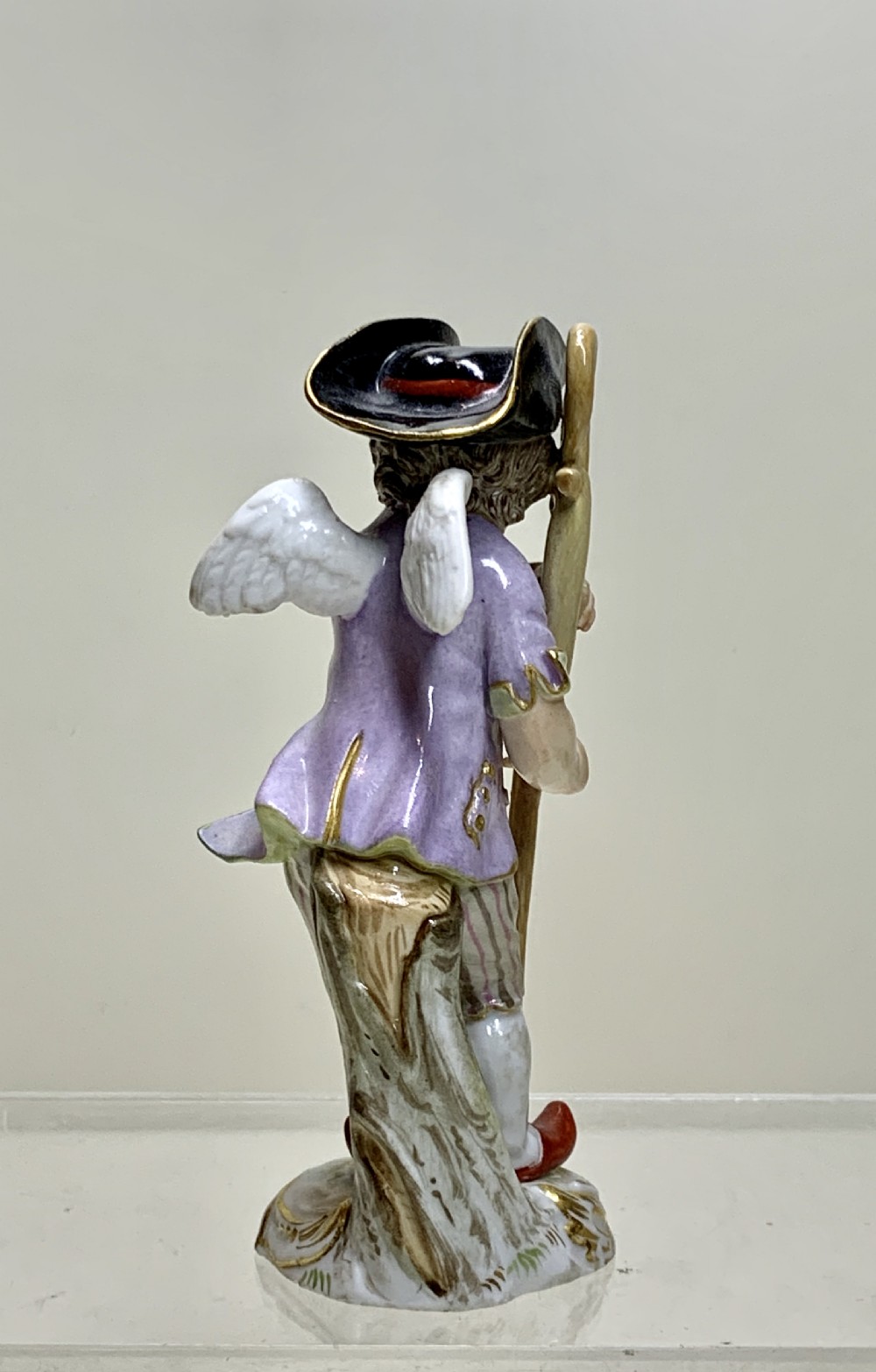 An 18th-century Meissen porcelain figurine depicting a winged Cupid disguised in a black tricorn hat, purple coat, and striped breeches resting on a staff. View V