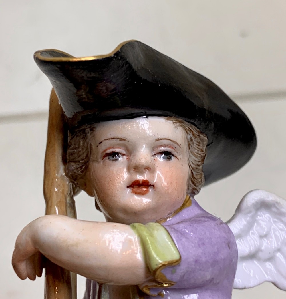 An 18th-century Meissen porcelain figurine depicting a winged Cupid disguised in a black tricorn hat, purple coat, and striped breeches resting on a staff. View XII
