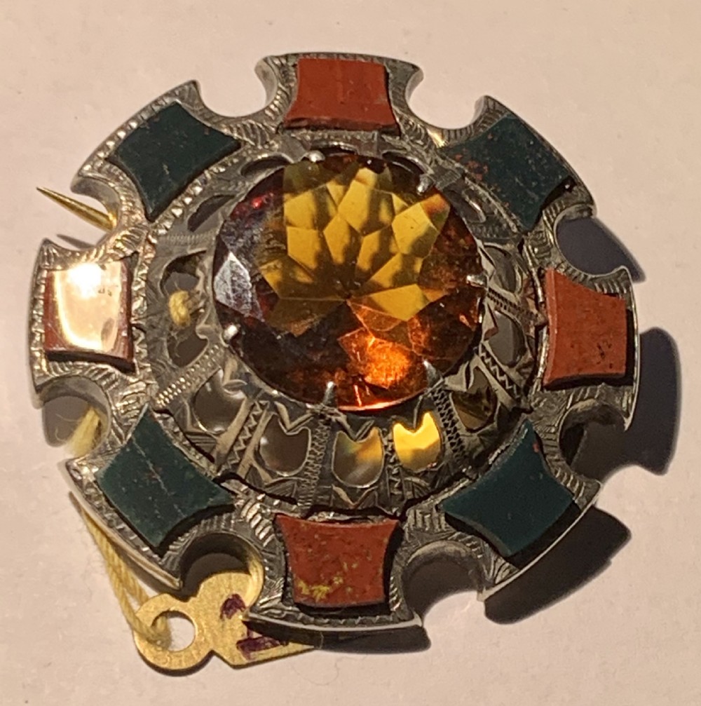 A circular Victorian silver Scottish brooch inlaid with red and green agate panels surrounding a large central faceted amber citrine stone.