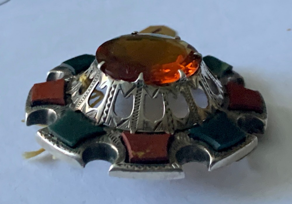A circular Victorian silver Scottish brooch inlaid with red and green agate panels surrounding a large central faceted amber citrine stone. View III