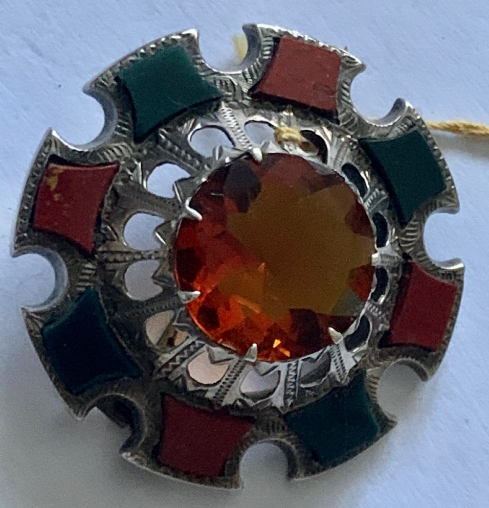 A circular Victorian silver Scottish brooch inlaid with red and green agate panels surrounding a large central faceted amber citrine stone. View IV