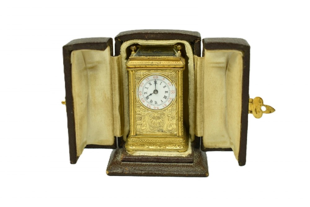 A miniature French gilt brass carriage clock with ornate scroll engraving and a white enamel dial, shown inside its open leather travelling case.