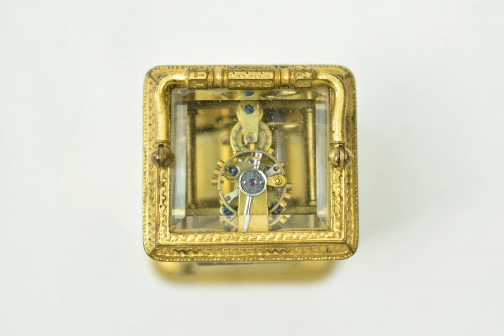 A miniature French gilt brass carriage clock with ornate scroll engraving and a white enamel dial, shown inside its open leather travelling case. View VI