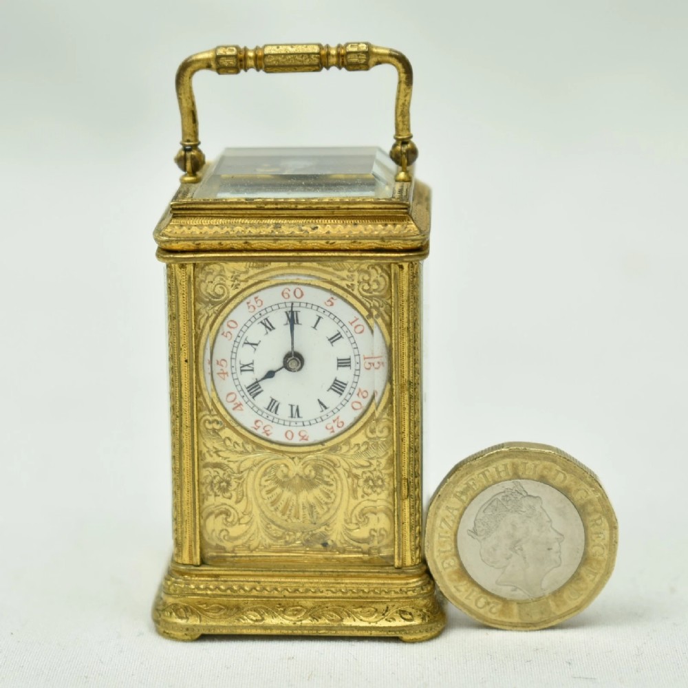 A miniature French gilt brass carriage clock with ornate scroll engraving and a white enamel dial, shown inside its open leather travelling case. View VIII