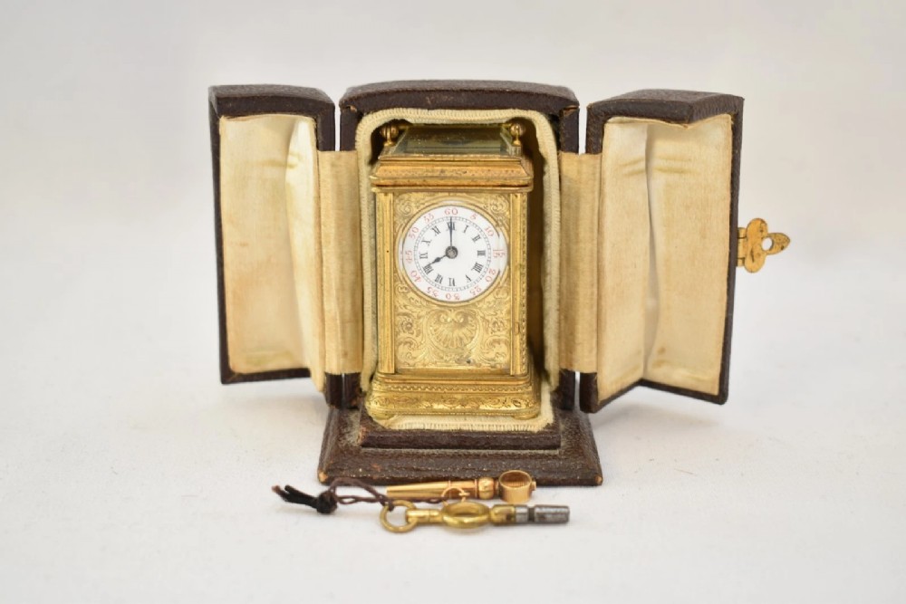 A miniature French gilt brass carriage clock with ornate scroll engraving and a white enamel dial, shown inside its open leather travelling case. View XII