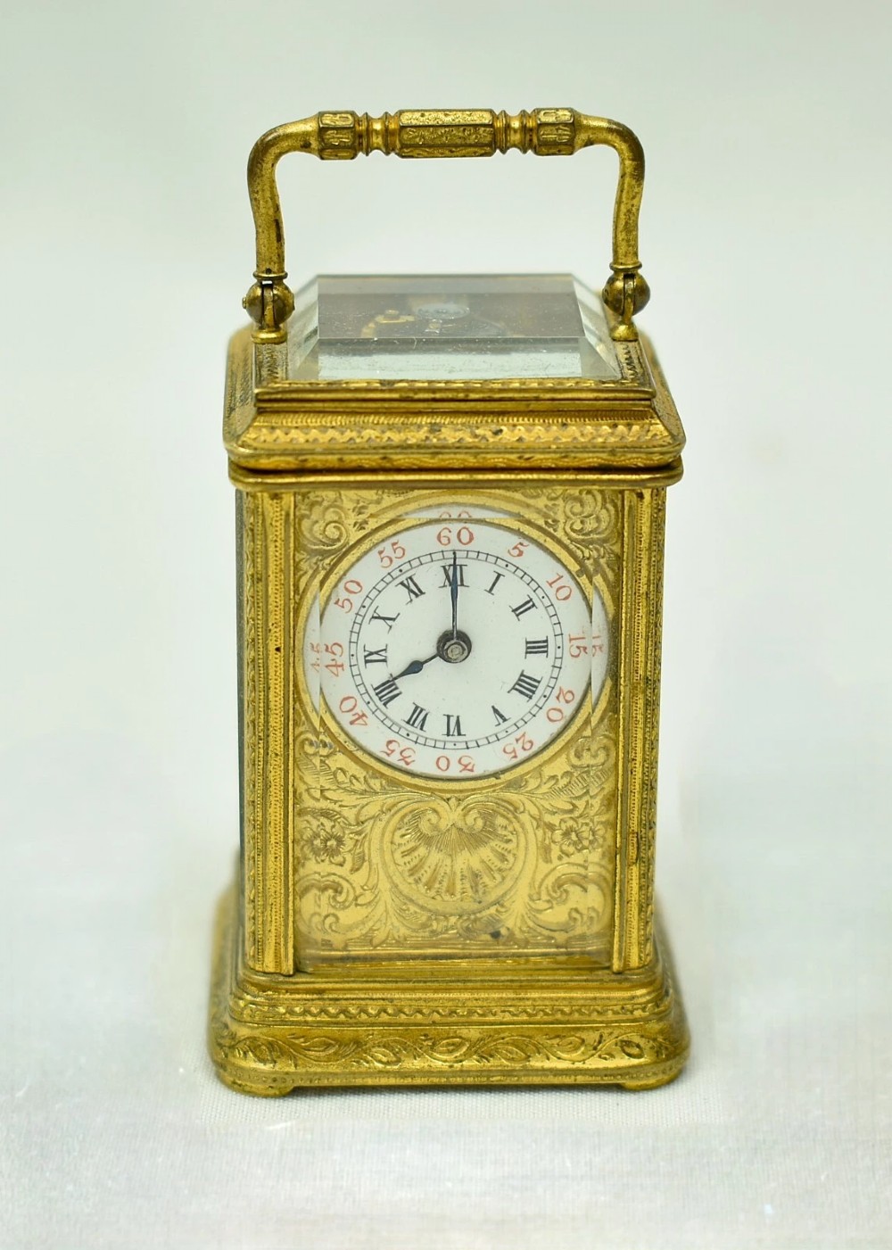 A miniature French gilt brass carriage clock with ornate scroll engraving and a white enamel dial, shown inside its open leather travelling case. View XIII