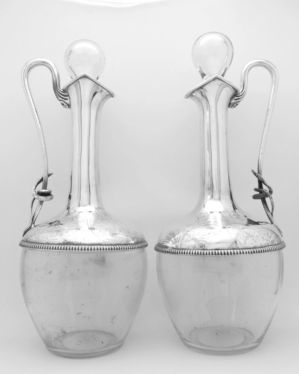 A pair of nineteenth-century clear crystal claret jugs featuring engraved silver mounts, tall necks, round stoppers, and silver handles shaped as entwined serpents.