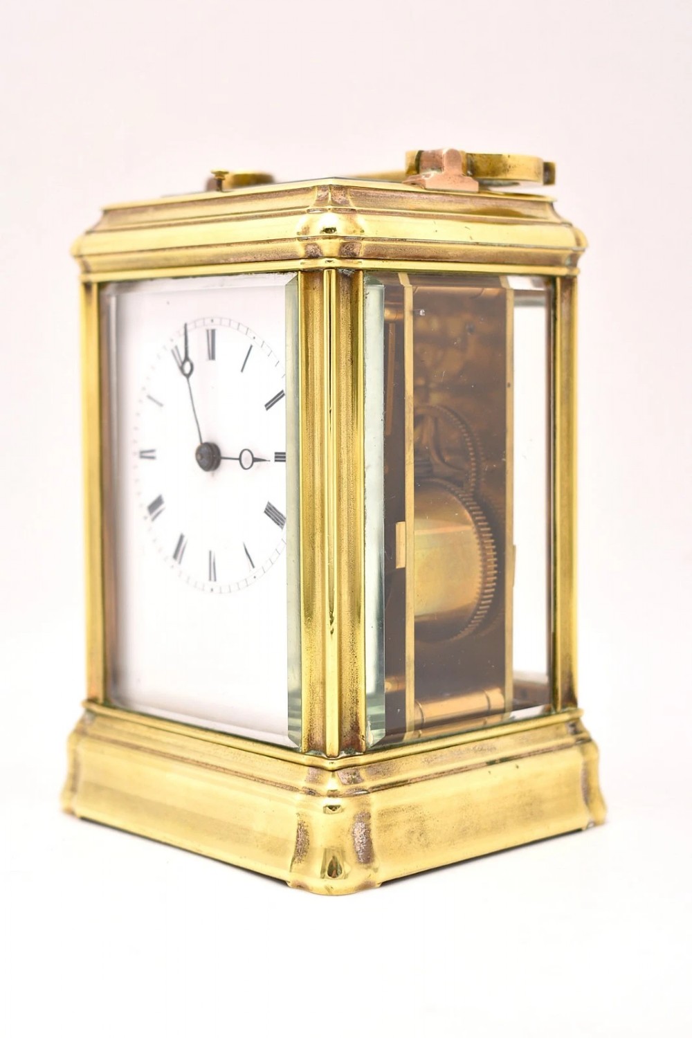 A rectangular polished brass French carriage clock featuring a doucine style case, top carrying handle, white enamel dial with Roman numerals, and glass side panels. View VII