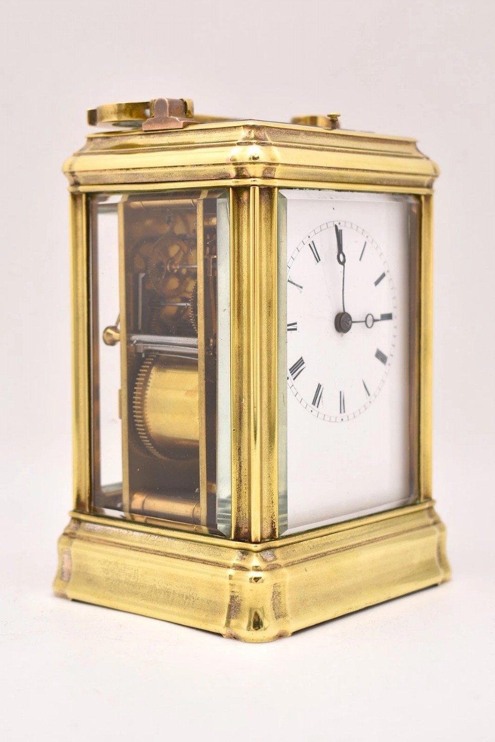 A rectangular polished brass French carriage clock featuring a doucine style case, top carrying handle, white enamel dial with Roman numerals, and glass side panels. View VIII