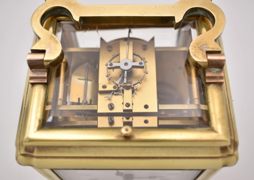 A rectangular polished brass French carriage clock featuring a doucine style case, top carrying handle, white enamel dial with Roman numerals, and glass side panels. View IX