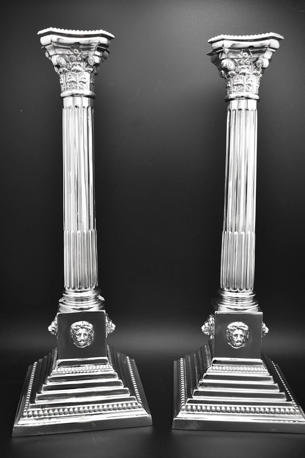 A pair of sterling silver candlesticks featuring fluted Corinthian columns, acanthus leaf capitals, lion mask motifs, and stepped square bases with beaded edging.