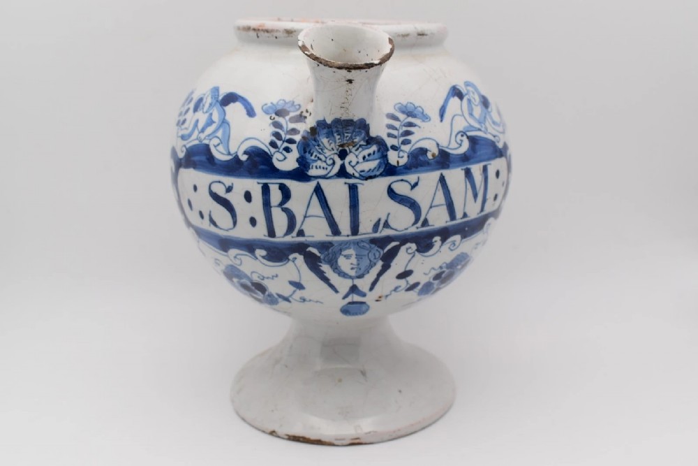 An 18th-century English delftware apothecary wet drug jar decorated in cobalt blue and white with a pouring spout, footed base, cherub motif, and the inscription S: BALSAM.
