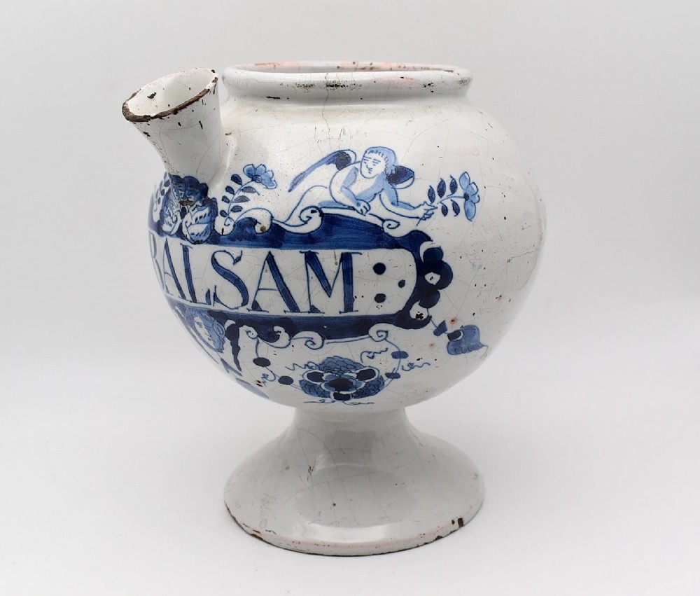 An 18th-century English delftware apothecary wet drug jar decorated in cobalt blue and white with a pouring spout, footed base, cherub motif, and the inscription S: BALSAM. View V