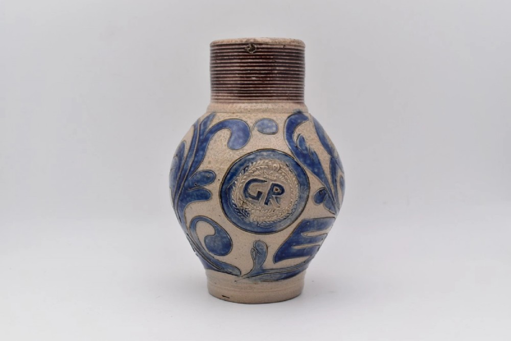 An 18th-century grey salt-glazed stoneware jug featuring cobalt blue floral scrollwork, a ribbed neck, and a central circular medallion stamped with the GR royal cipher.