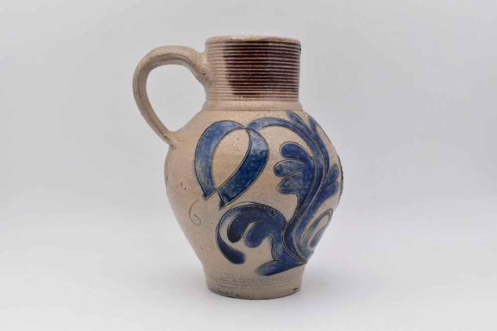 An 18th-century grey salt-glazed stoneware jug featuring cobalt blue floral scrollwork, a ribbed neck, and a central circular medallion stamped with the GR royal cipher. View II