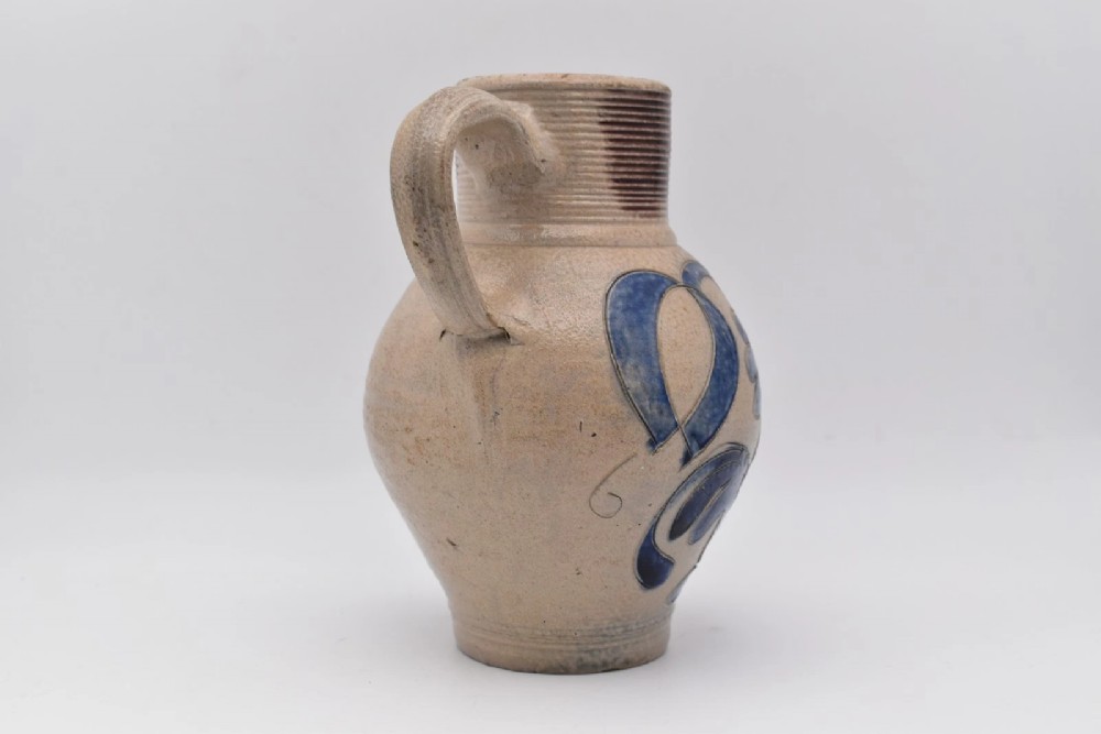 An 18th-century grey salt-glazed stoneware jug featuring cobalt blue floral scrollwork, a ribbed neck, and a central circular medallion stamped with the GR royal cipher. View III