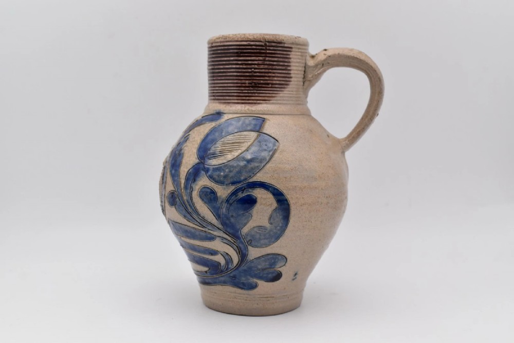 An 18th-century grey salt-glazed stoneware jug featuring cobalt blue floral scrollwork, a ribbed neck, and a central circular medallion stamped with the GR royal cipher. View IV
