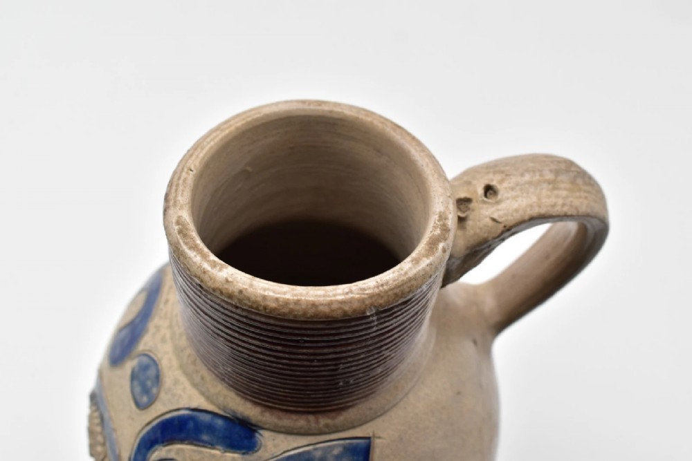 An 18th-century grey salt-glazed stoneware jug featuring cobalt blue floral scrollwork, a ribbed neck, and a central circular medallion stamped with the GR royal cipher. View V