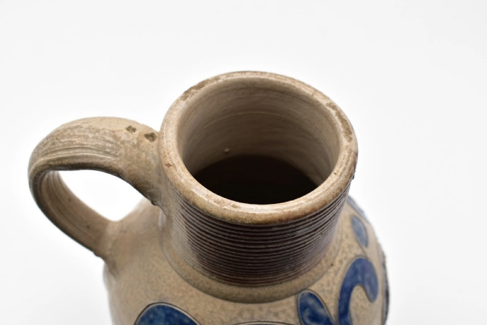 An 18th-century grey salt-glazed stoneware jug featuring cobalt blue floral scrollwork, a ribbed neck, and a central circular medallion stamped with the GR royal cipher. View VI