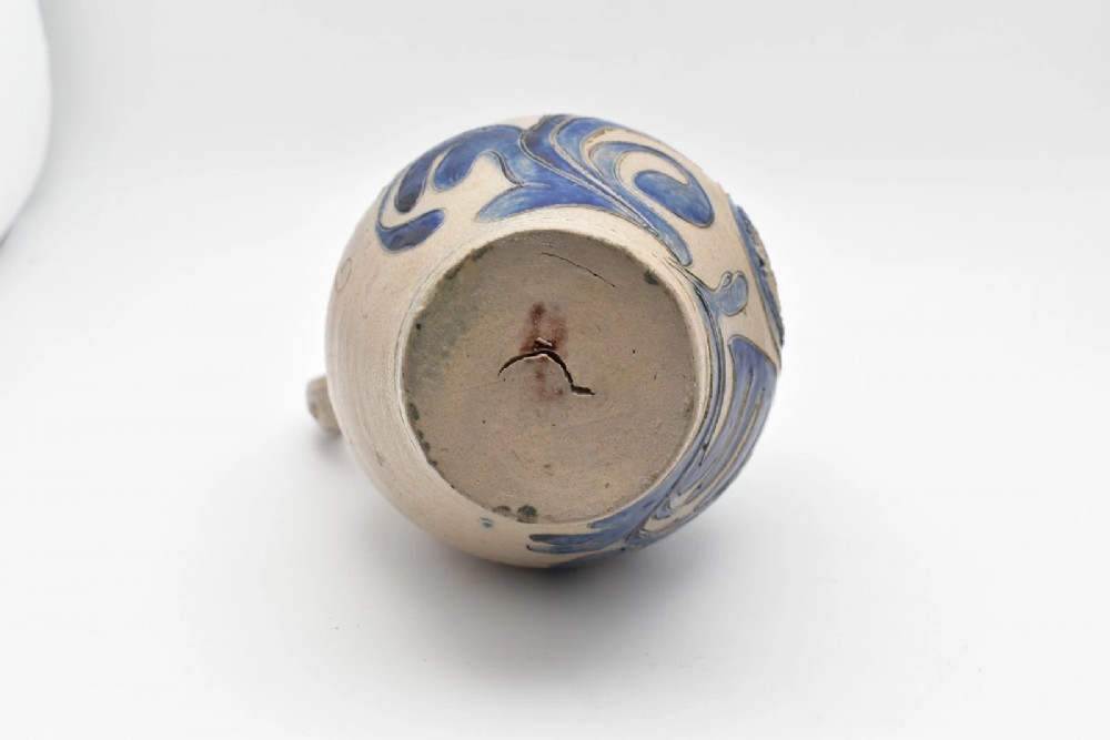 An 18th-century grey salt-glazed stoneware jug featuring cobalt blue floral scrollwork, a ribbed neck, and a central circular medallion stamped with the GR royal cipher. View VII