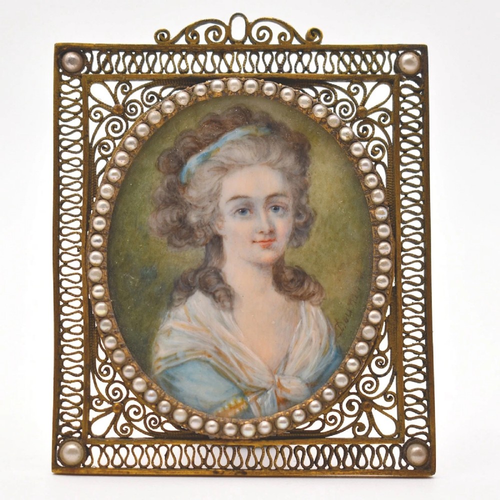 An 18th-century oval watercolour portrait miniature depicting a woman with curled hair, housed within a seed pearl bezel and a gilt filigree rectangular frame.