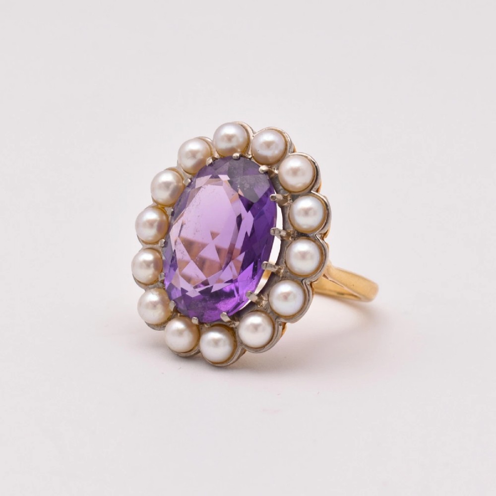 An 18ct yellow gold cluster ring featuring a centrally set oval faceted purple amethyst surrounded by a halo of fourteen small white pearls. View VI