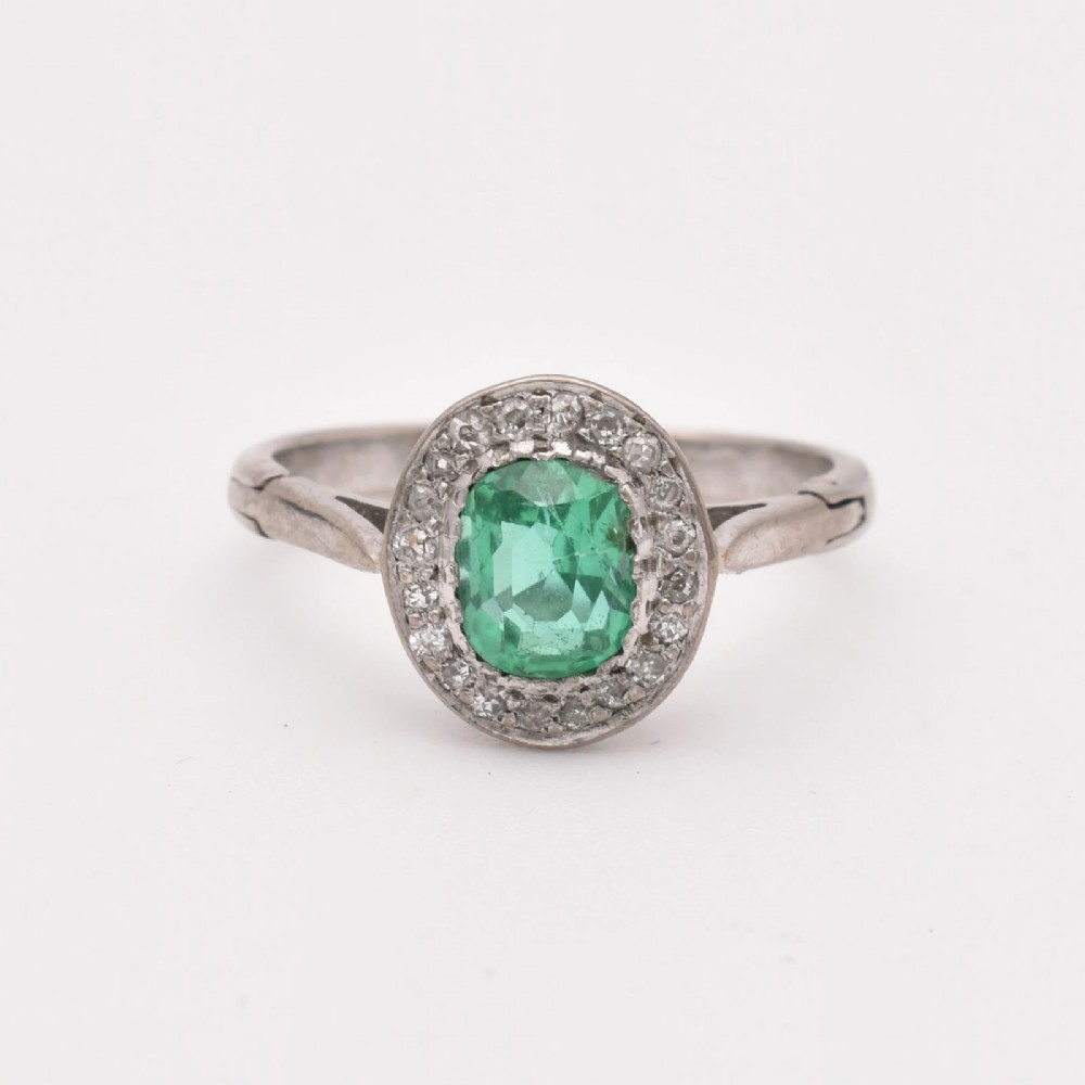 An oval green emerald set at the centre of a white gold cluster ring surrounded by small diamonds with an adjustable band.