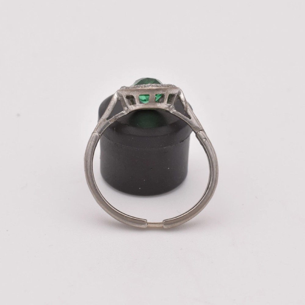 An oval green emerald set at the centre of a white gold cluster ring surrounded by small diamonds with an adjustable band. View II