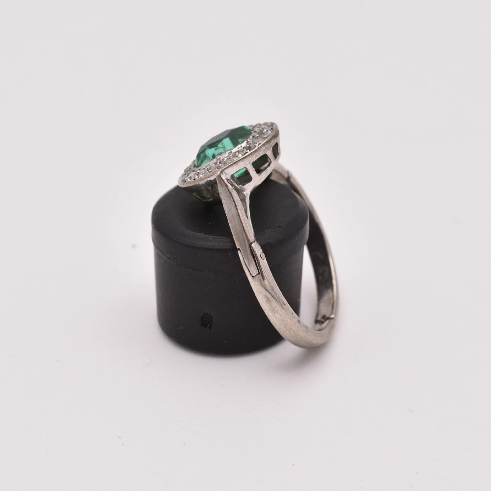 An oval green emerald set at the centre of a white gold cluster ring surrounded by small diamonds with an adjustable band. View IV