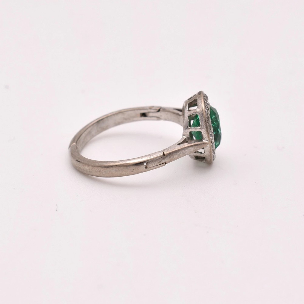 An oval green emerald set at the centre of a white gold cluster ring surrounded by small diamonds with an adjustable band. View V