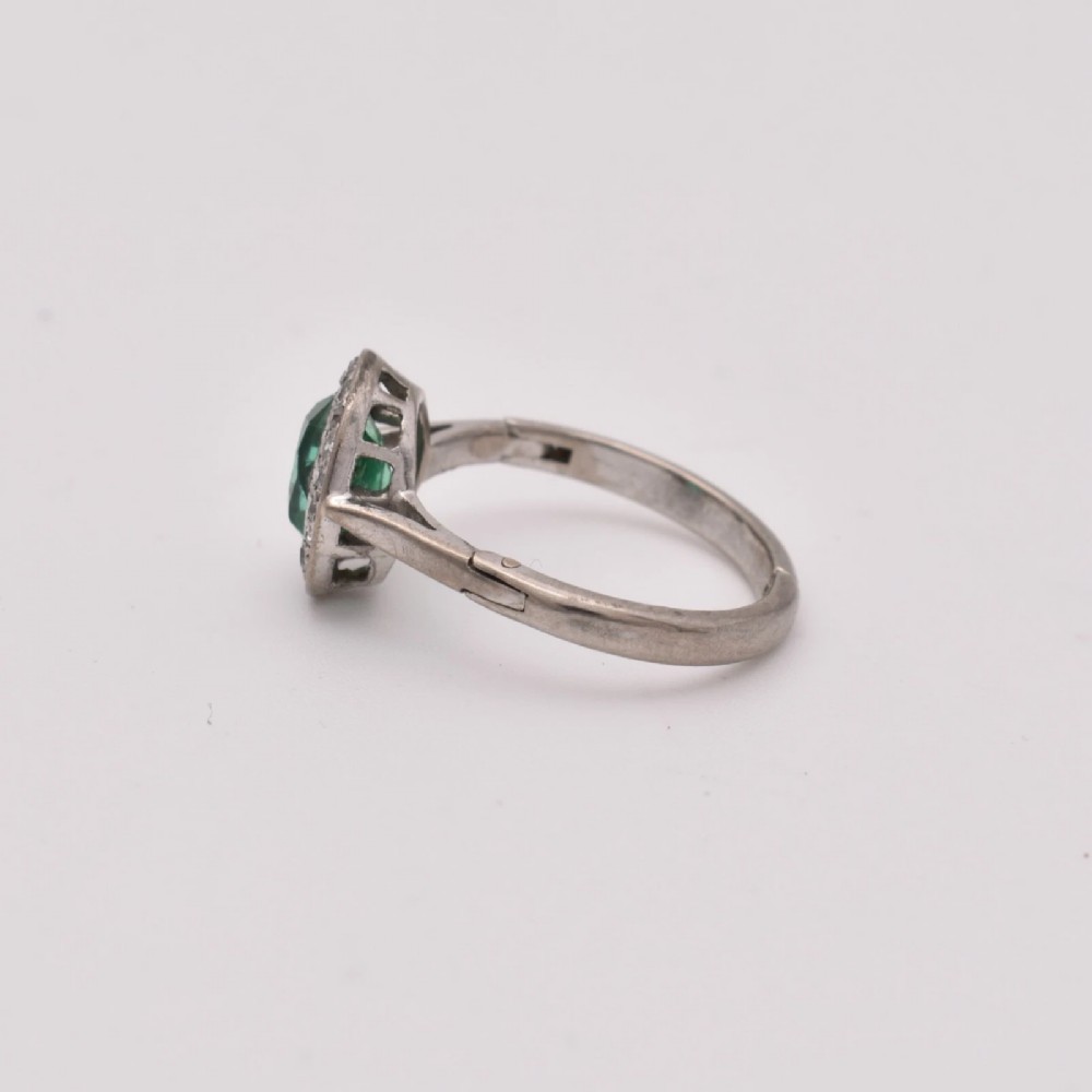 An oval green emerald set at the centre of a white gold cluster ring surrounded by small diamonds with an adjustable band. View VI