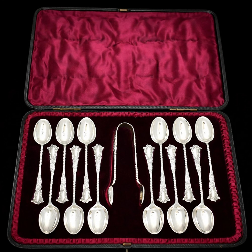 A set of twelve silver teaspoons with twisted stems and matching sugar tongs arranged inside a fitted burgundy velvet-lined case.