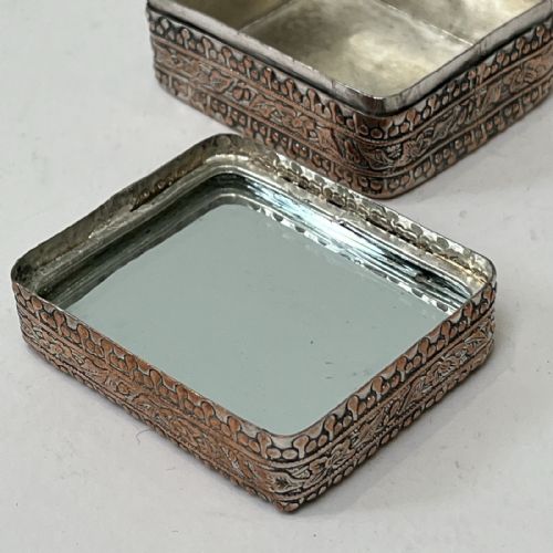 fig 2. A small rectangular silver-plated metal pill box with intricately chased foliate sides and a hand-painted enamel lid depicting a figure in green traditional dress.