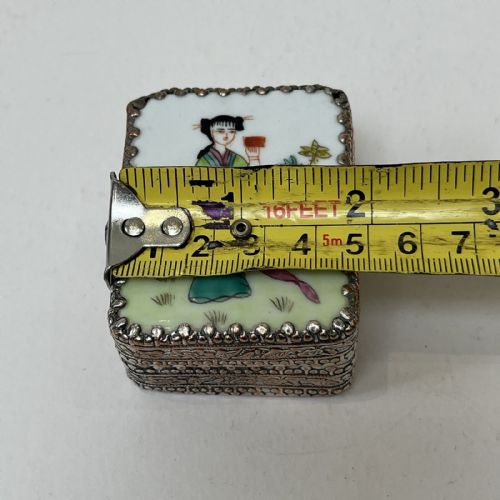 fig 3. A small rectangular silver-plated metal pill box with intricately chased foliate sides and a hand-painted enamel lid depicting a figure in green traditional dress.