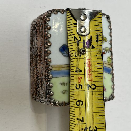 fig 4. A small rectangular silver-plated metal pill box with intricately chased foliate sides and a hand-painted enamel lid depicting a figure in green traditional dress.