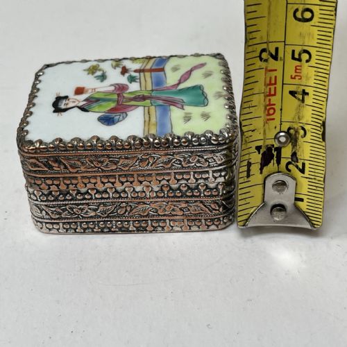 fig 5. A small rectangular silver-plated metal pill box with intricately chased foliate sides and a hand-painted enamel lid depicting a figure in green traditional dress.