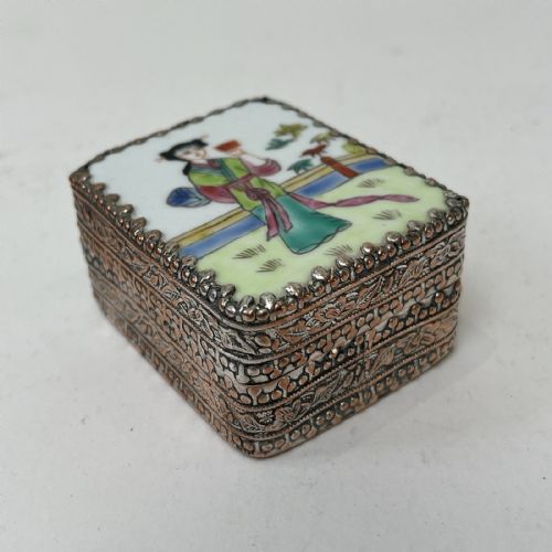 fig 6. A small rectangular silver-plated metal pill box with intricately chased foliate sides and a hand-painted enamel lid depicting a figure in green traditional dress.