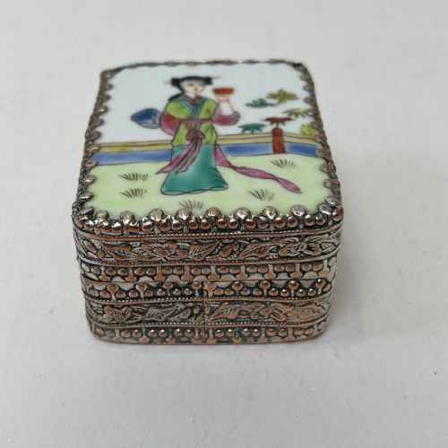 fig 7. A small rectangular silver-plated metal pill box with intricately chased foliate sides and a hand-painted enamel lid depicting a figure in green traditional dress.