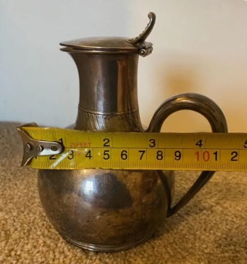 fig 6. A small bulbous silver-plated pitcher featuring a hinged lid, thumbpiece, curved handle, and a ribbed decorative collar.