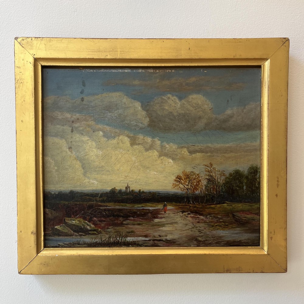 A 19th-century oil painting featuring a figure walking along a dirt pathway towards a distant church spire under a sky filled with clouds, set within a wide gold frame.
