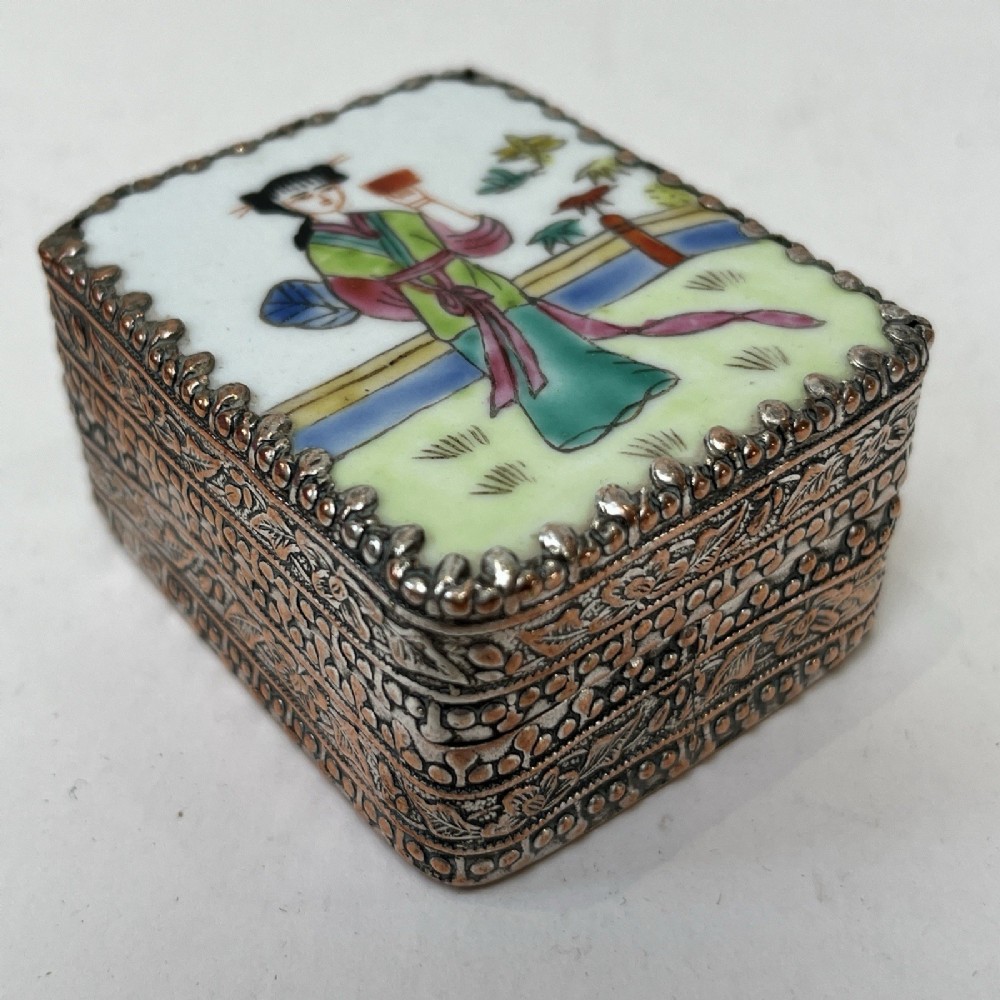 A small rectangular silver-plated metal pill box with intricately chased foliate sides and a hand-painted enamel lid depicting a figure in green traditional dress.