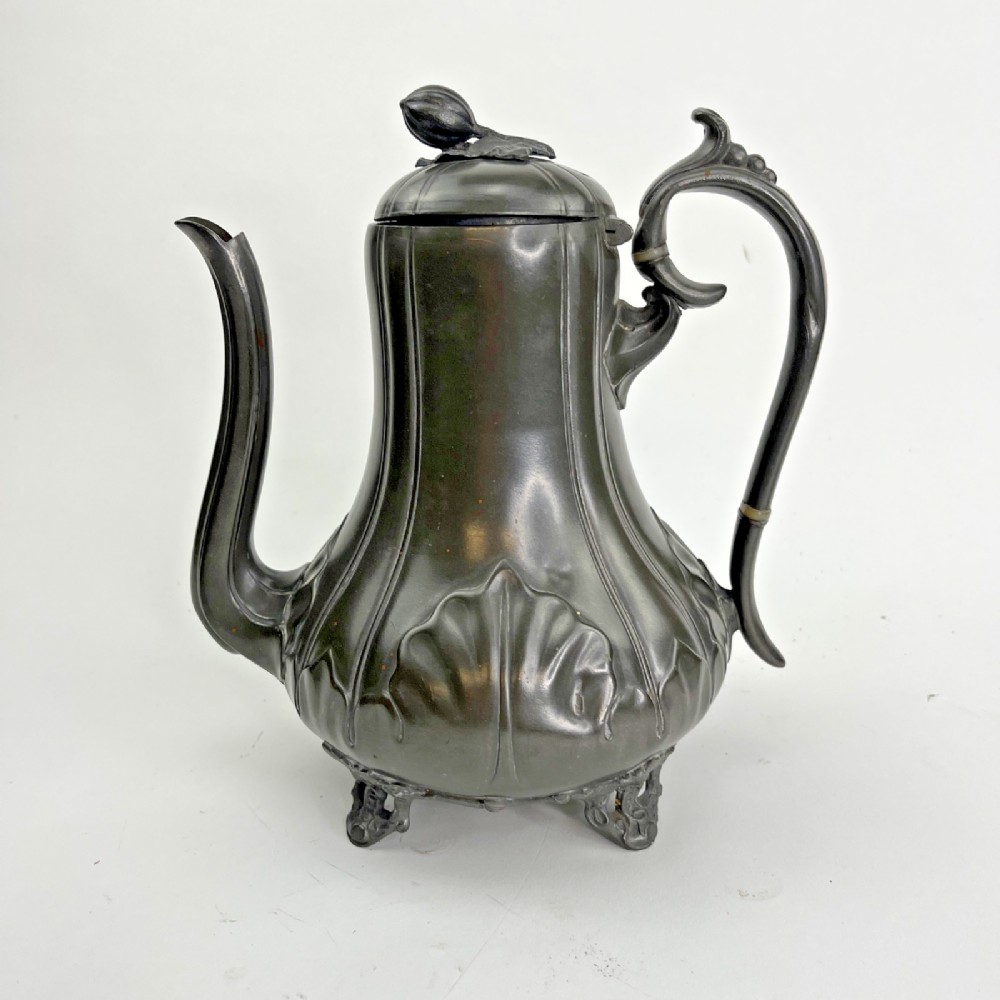 A dark grey Victorian Britannia metal hot water pot featuring a high gooseneck spout, leaf-lobed body, scroll handle, and floral bud finial.