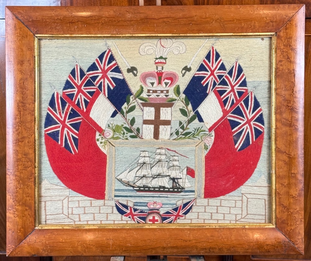 A 19th-century sailors woolwork picture featuring a sailing ship surrounded by Union flags and heraldic motifs, set within a birds eye maple frame.
