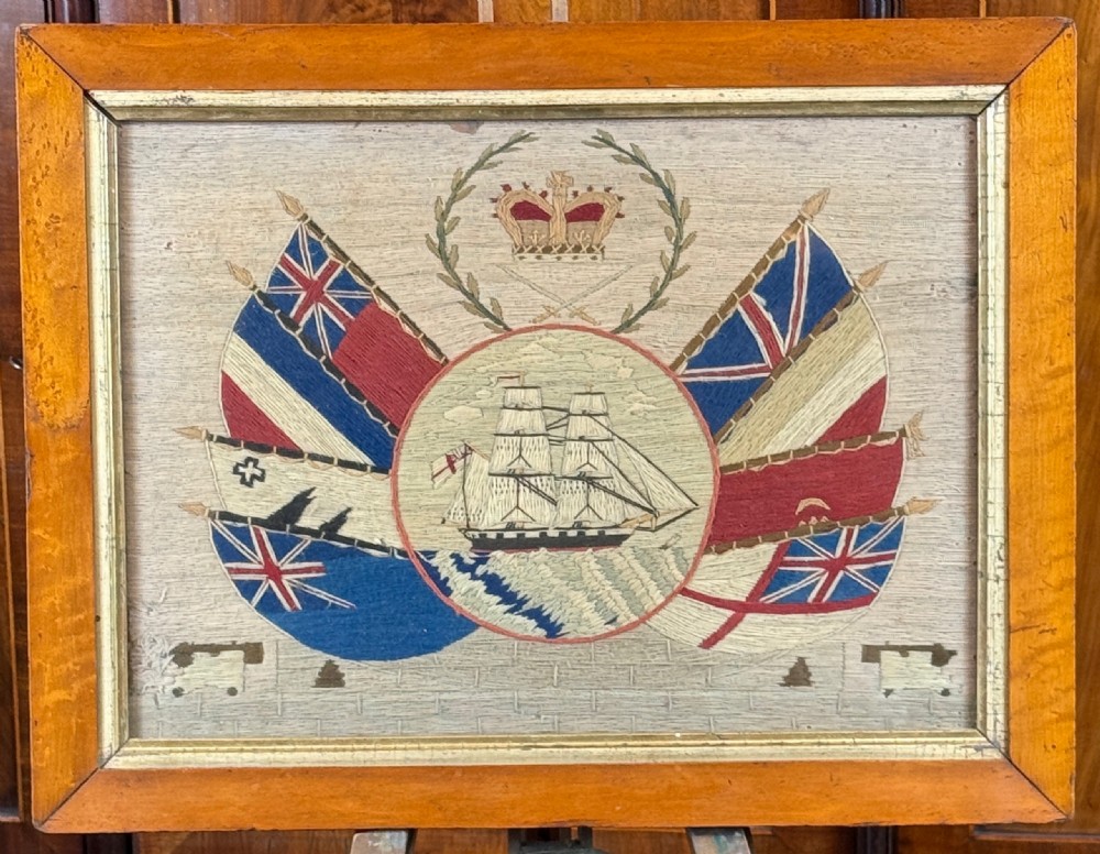A framed 19th-century sailors wool embroidery featuring a central sailing ship surrounded by flags, a crown, laurel branches, and miniature cannons.