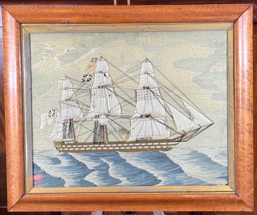 A framed 19th-century wool needlework depicting a three-masted sailing ship on blue embroidered waves set within a wooden frame.