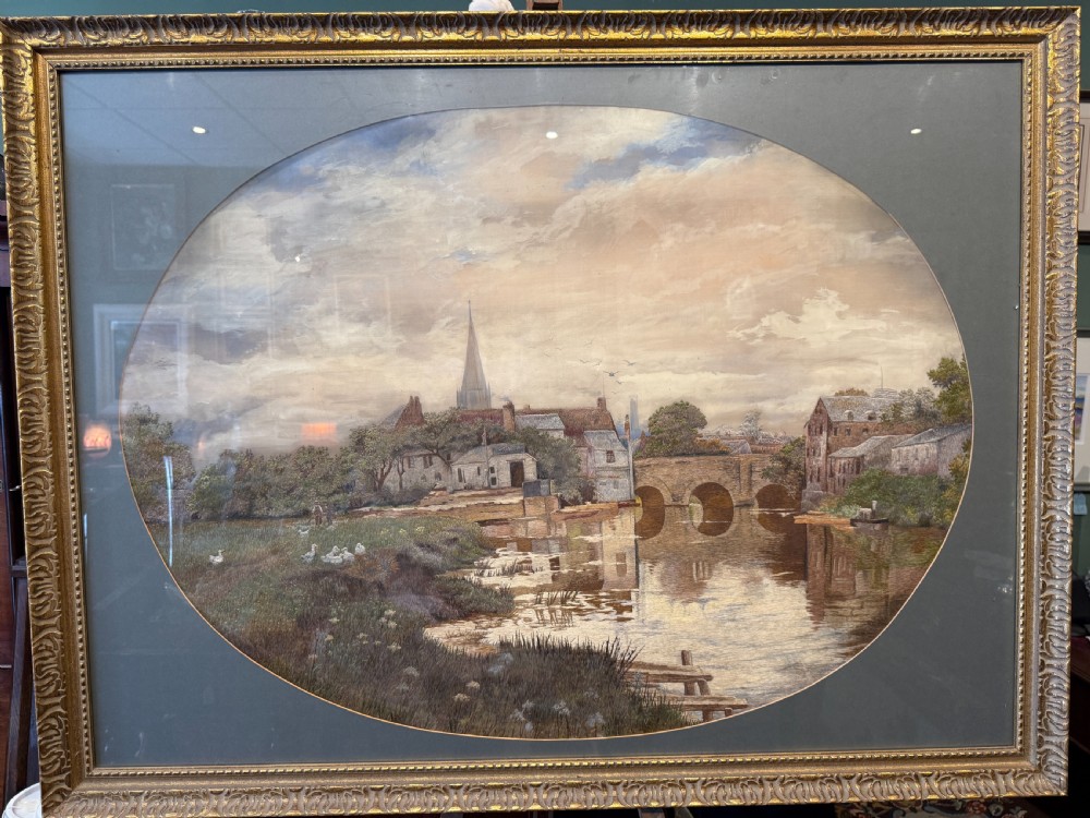 A large Victorian embroidery and watercolour landscape depicting a town and arched bridge over a river, set in a gold gilt frame with an oval grey mount.