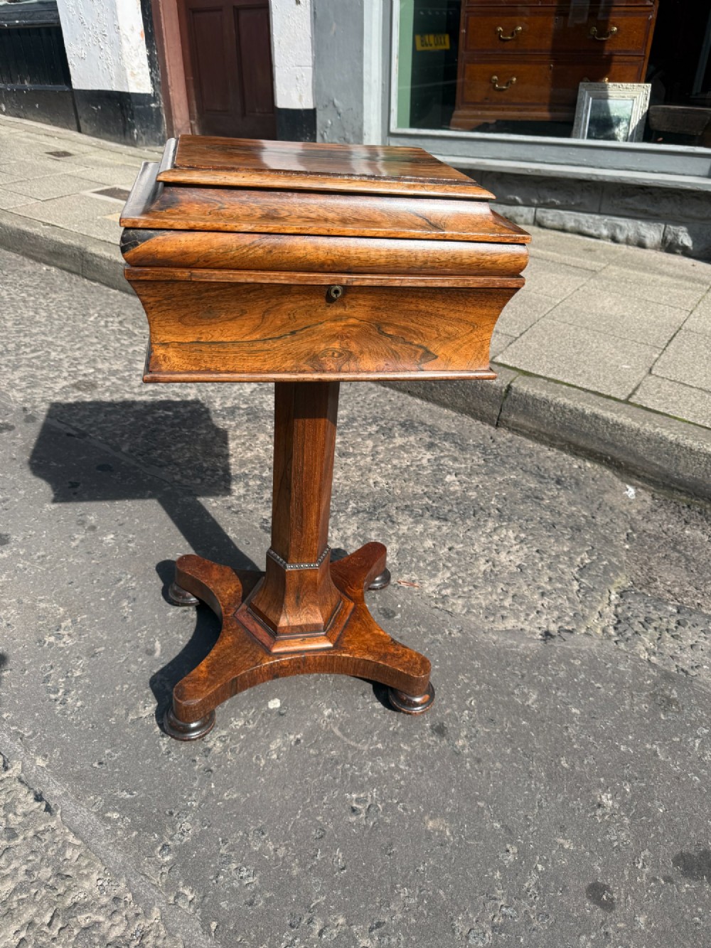 A Regency rosewood teapoy featuring a sarcophagus-shaped hinged lid, faceted pedestal column, and quadripartite plinth base on bun feet.