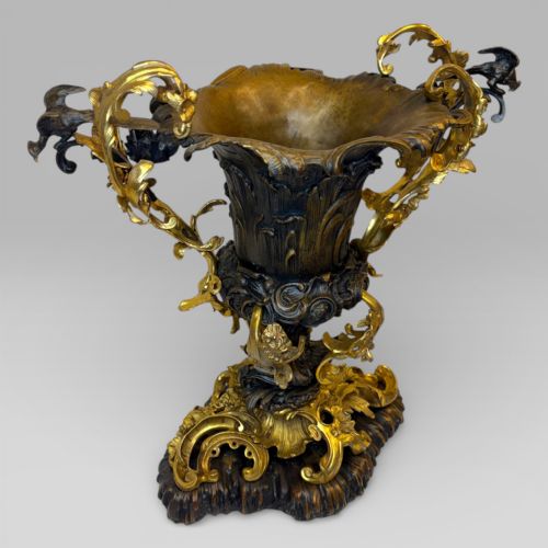 fig 1. An ornate gilt and patinated bronze centrepiece urn featuring twin scrolled foliate handles, a scalloped rim, and a sculpted rocaille base.
