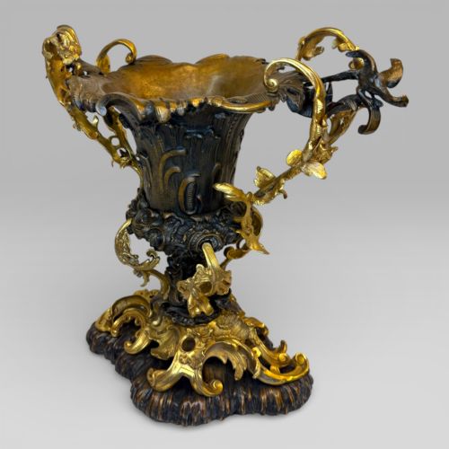 fig 2. An ornate gilt and patinated bronze centrepiece urn featuring twin scrolled foliate handles, a scalloped rim, and a sculpted rocaille base.