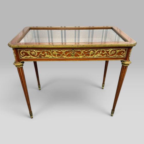 fig 1. A rectangular French Louis XVI style bijouterie display table featuring a glazed glass top, decorative gilt bronze mounts along the frieze, and slender tapered legs ending in brass sabots.